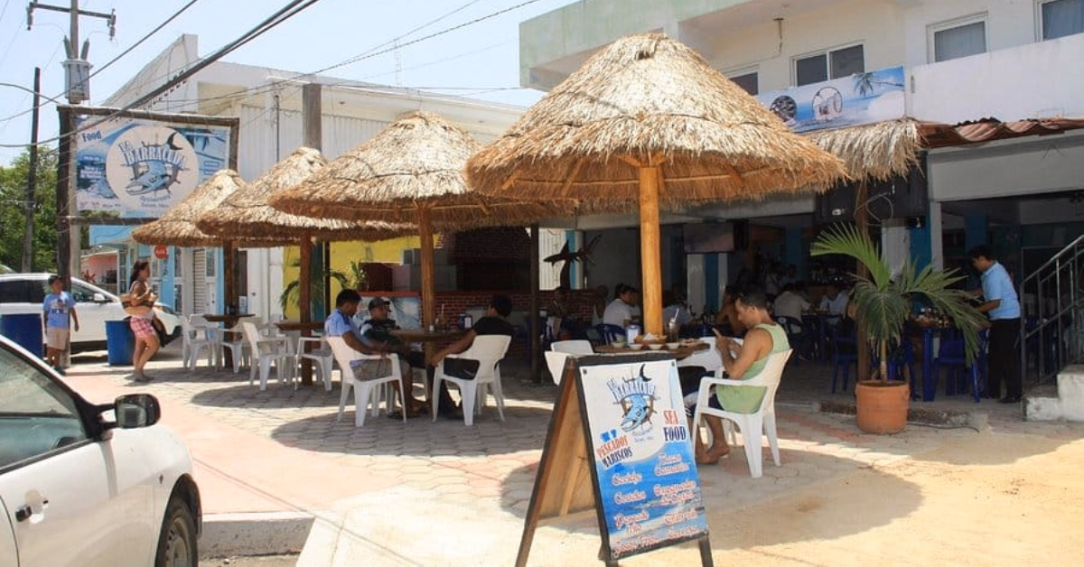 Outdoor seafood restaurant with thatched umbrellas, white plastic tables and chairs, and people dining. A menu board stands at the entrance, marked as one of the top Seafood Restaurants in Tulum. Nearby buildings and a parked car are visible.