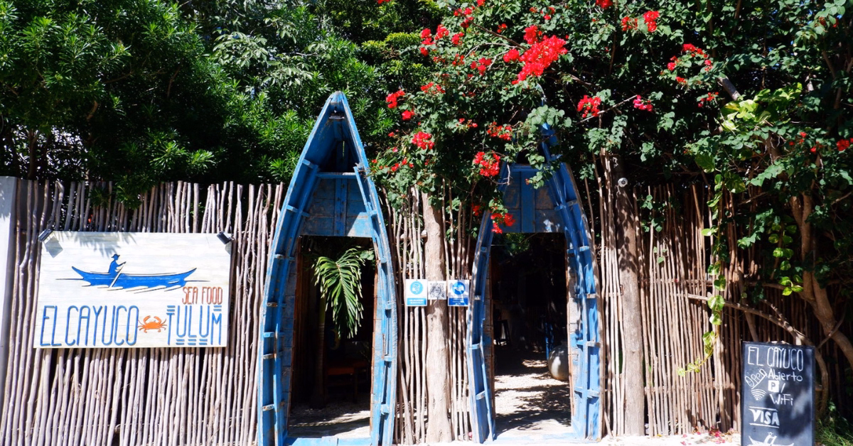 Entrance to a rustic restaurant with two blue boat frames as doorways, surrounded by a wooden fence, lush greenery, and vibrant red flowers. Signs display the name El Cayuco Tulum, one of the standout seafood restaurants in Tulum.