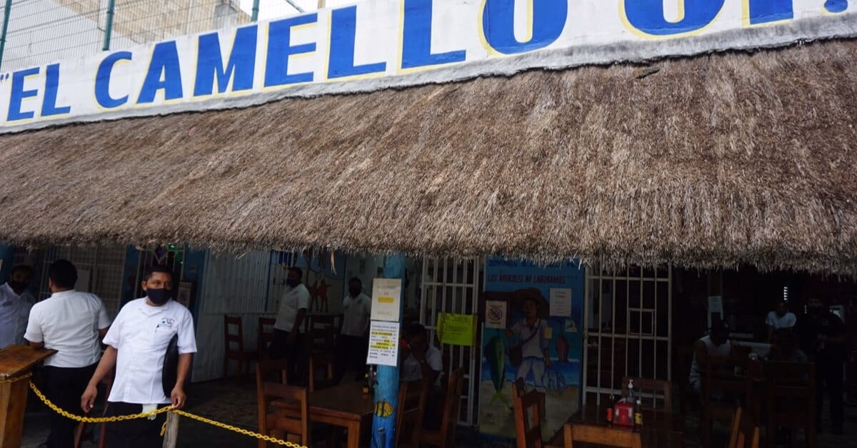 A thatched-roof restaurant exterior with the sign El Camello partially visible. Several staff in white shirts and face masks stand outside—one of the popular seafood restaurants in Tulum, with tables and chairs seen inside under the roof.