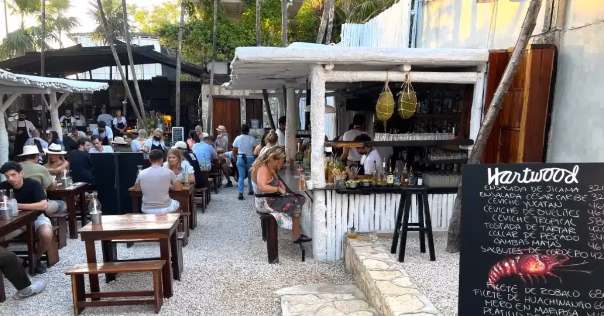 Open-air restaurant with wooden tables and benches, people dining, and a white rustic bar. A handwritten chalkboard menu stands near the bar, and tropical trees are visible—perfect for enjoying Seafood Restaurants in Tulum’s lively, relaxed ambiance.