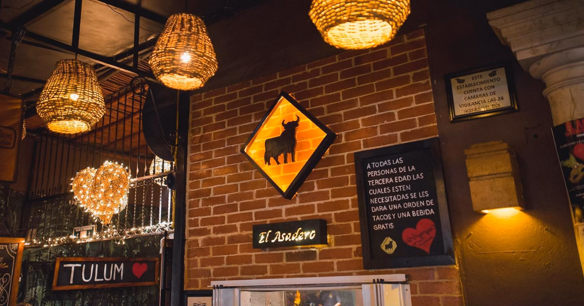 Warmly lit steak restaurant interior with wicker hanging lamps, a glowing heart-shaped light, and a brick wall featuring a yellow bull sign. A “Tulum” sign hints at one of the top Steak Restaurants in Tulum, with string lights adding to the charm.