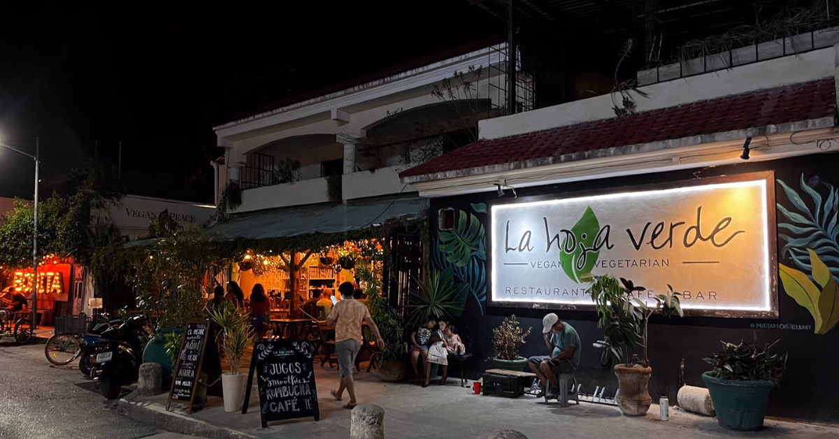 Nighttime street view of La Hoja Verde, one of the top Vegan Vegetarian Restaurants in Tulum, featuring outdoor seating, potted plants, an illuminated sign, and people relaxing or strolling near the entrance.