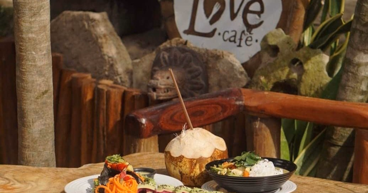 A rustic outdoor table displays fresh dishes, a coconut drink, and colorful vegetables. In the background, a wooden sign reads Love Cafe amidst greenery—a charming spot among Vegan Vegetarian Restaurants in Tulum.