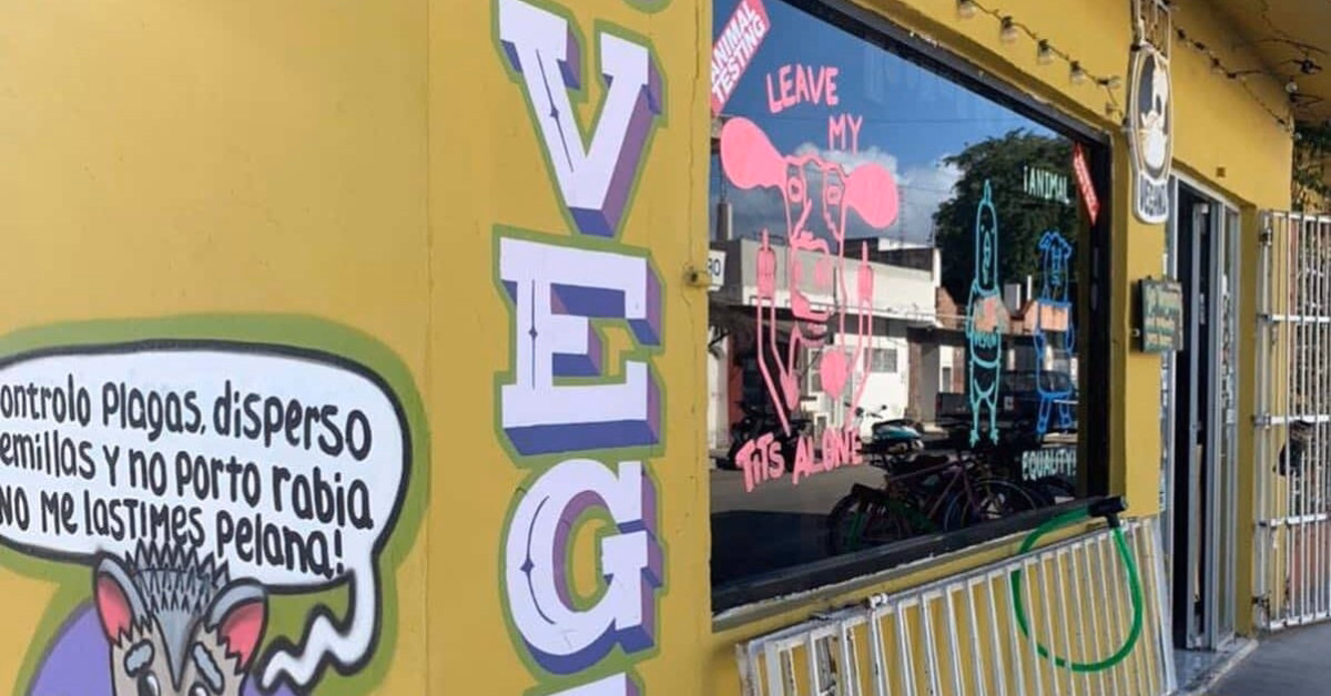 A yellow building with colorful signs, including a cartoon rat and large VEG letters, stands out among Vegan Vegetarian Restaurants in Tulum. The window displays playful rat drawings and quirky phrases. A bike rack sits out front.