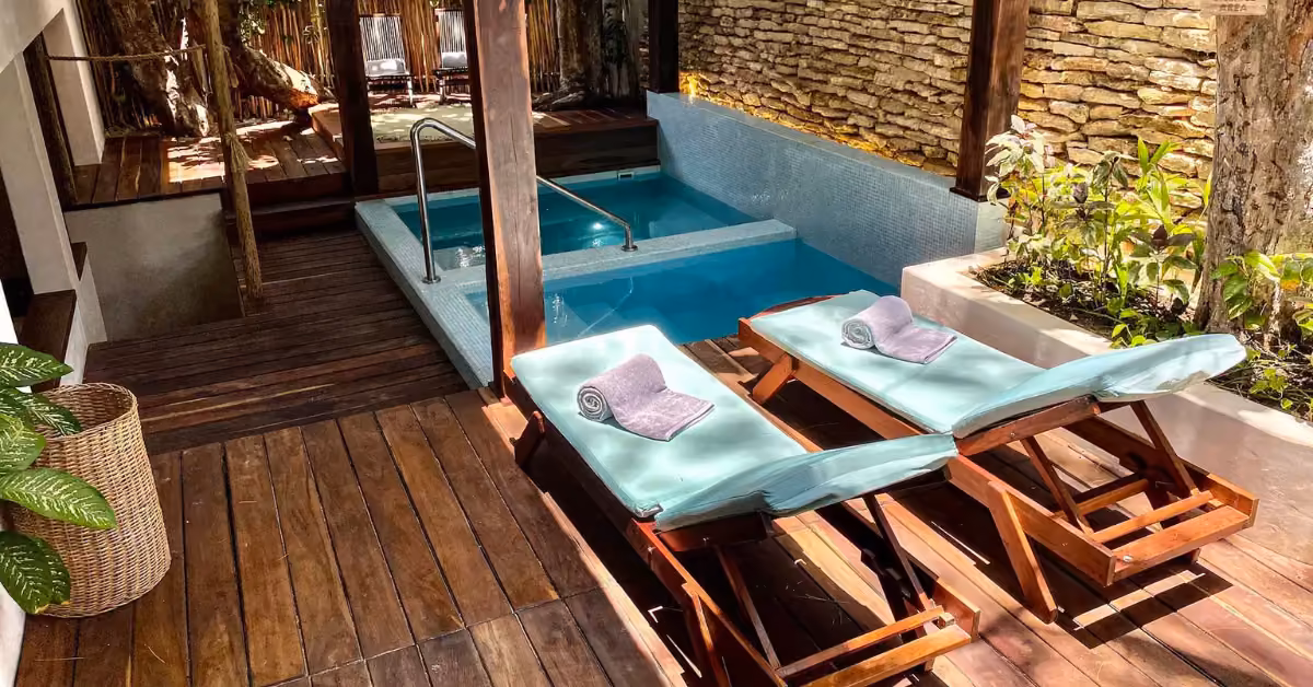 Two lounge chairs with towels face a private pool on wooden decking, surrounded by lush plants and a stone wall for relaxation.