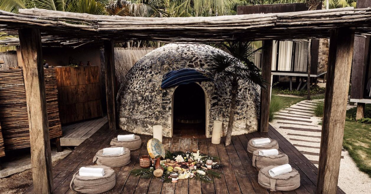A rustic stone temazcal sweat lodge sits on a wooden deck under a pergola, surrounded by tropical plants—an inviting space for Temazcal Ceremonies in Tulum. Cushions, towels, ceremonial objects, and food are beautifully arranged in the center.