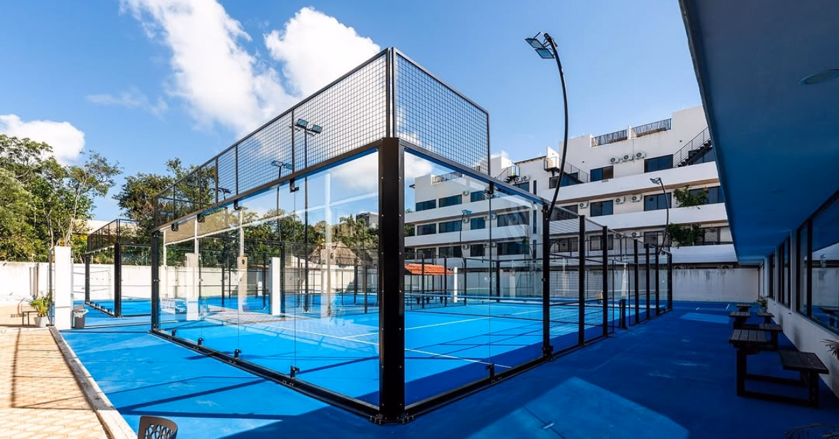 Blue padel court with glass walls, benches, and modern buildings nearby under a sunny sky with scattered clouds.