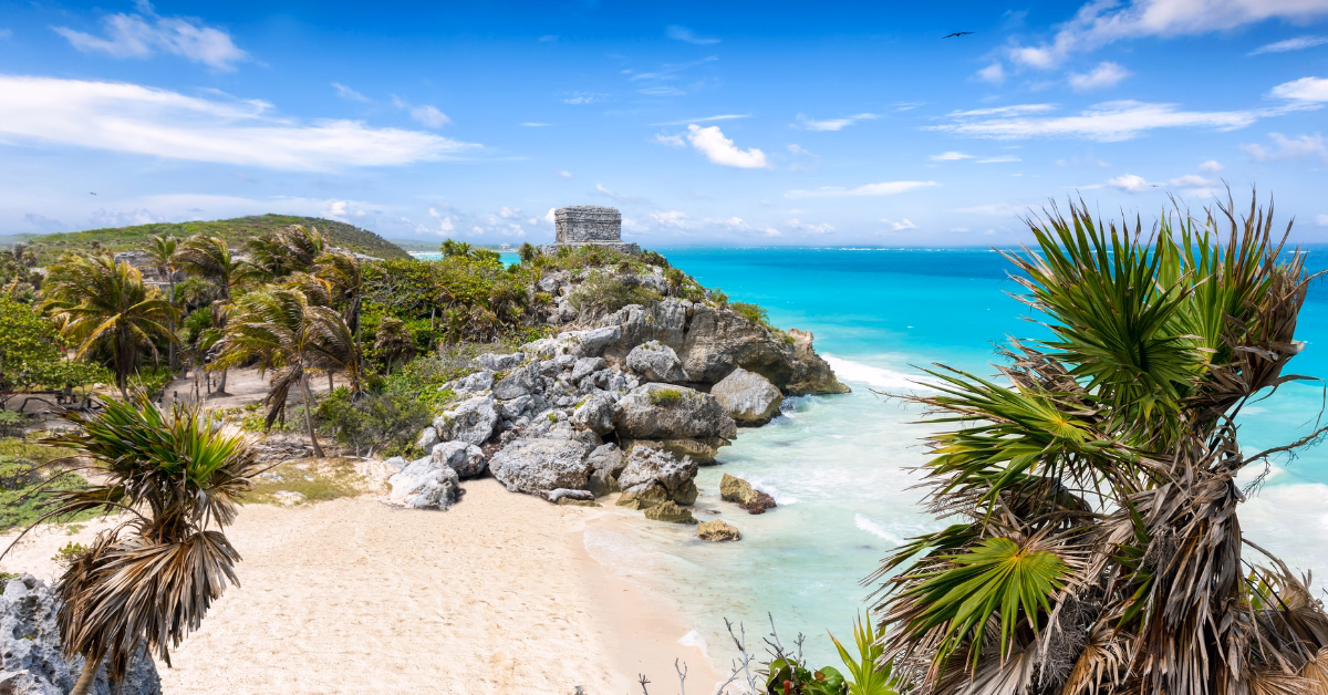 A sandy beach with turquoise water, rocky cliffs, and palm trees. Tulum’s Archaeological Site, an ancient stone ruin, sits atop the cliff under a blue sky with scattered clouds in Tulum, Mexico.