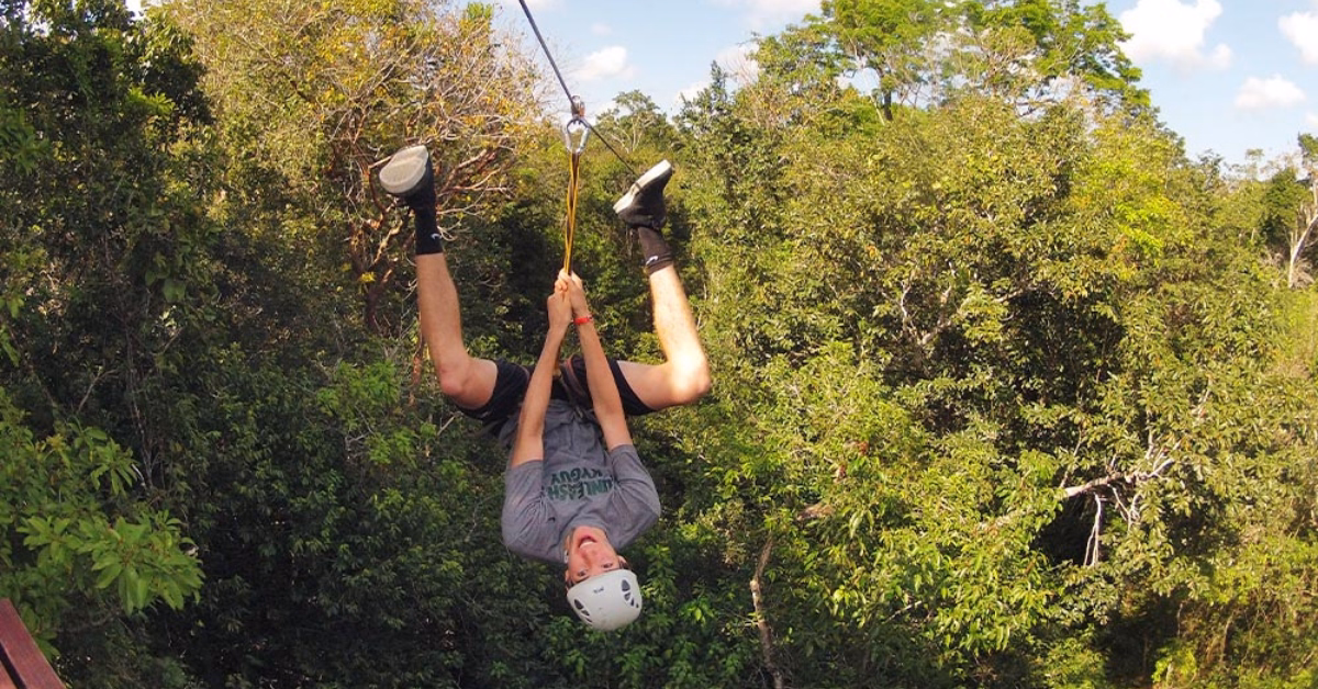 A helmeted person hangs upside down on a zipline above green trees, smiling with legs hooked over the cable in Tulum.