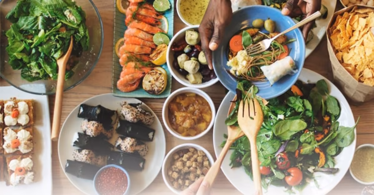 Friends reaching for shared plates of salads, sushi, shrimp, chips, and wraps on a lively table filled with fresh dishes.