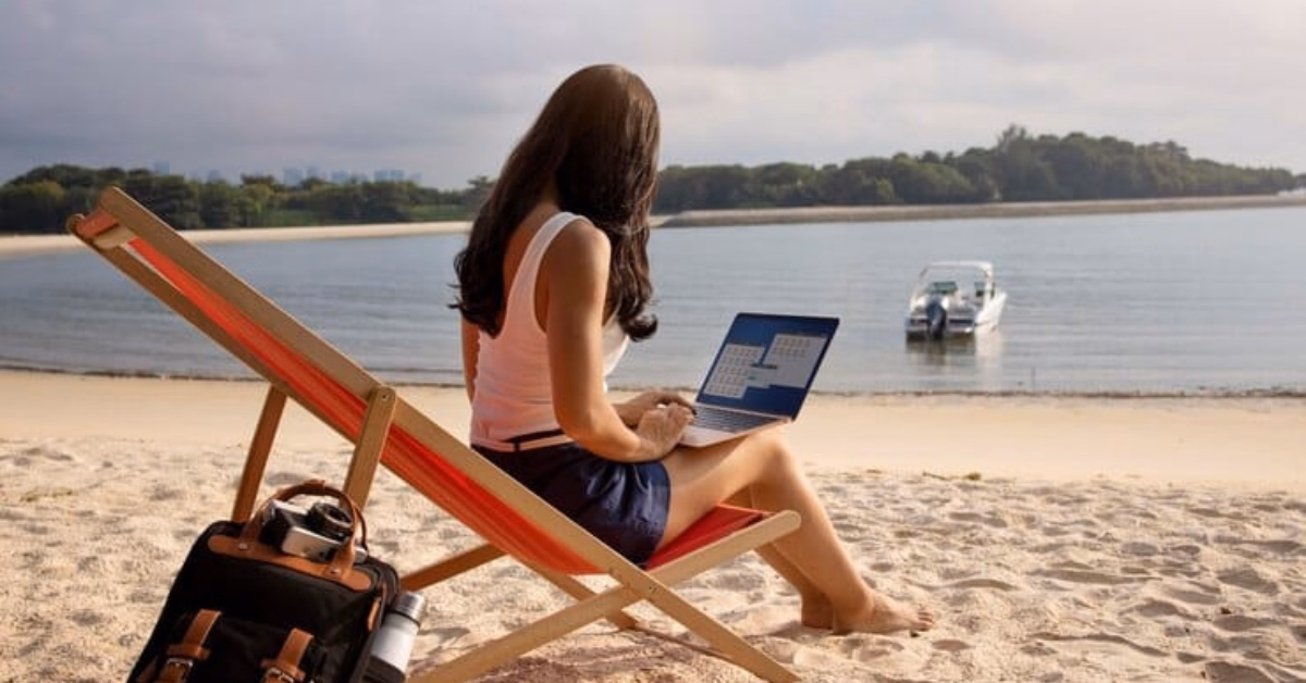 A woman in a beach chair works on her laptop by the water. A boat, backpack, trees, and distant buildings are nearby under clouds.