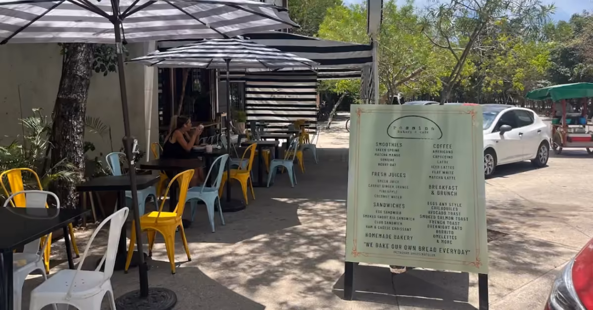 Outdoor café with yellow and blue chairs, striped umbrellas, a menu sign, trees, greenery, and a parked white car in the background.