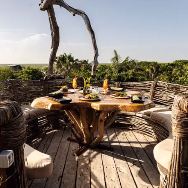 A rustic outdoor wooden table set with food and drinks sits on a deck at Kanan Restaurant, surrounded by woven branch railings, cushioned seats, and lush greenery under a clear sky.