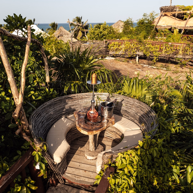 A cozy round seating area with a woven wooden backrest, cushioned benches, and a small table with a decanter and glasses at Kanan Restaurant, surrounded by lush green plants and overlooking the ocean in the distance.