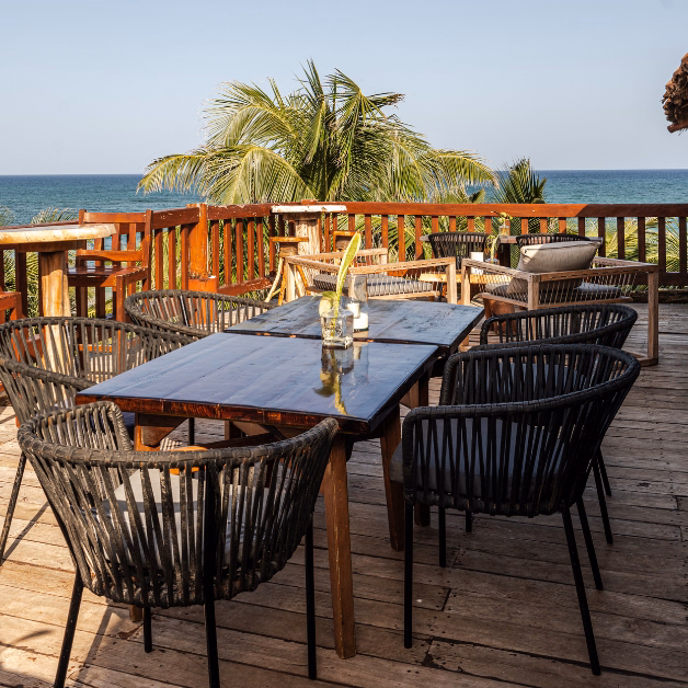 Outdoor wooden deck at Kanan Restaurant with black woven chairs and tables overlooking the ocean, surrounded by palm trees under a clear blue sky. A glass vase with a single flower decorates one table.