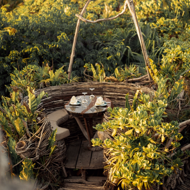 A round wooden dining table set for two with napkins and glasses sits inside a nest-like structure at Karma Rooftop, surrounded by lush green plants and trees, creating an intimate outdoor dining atmosphere.