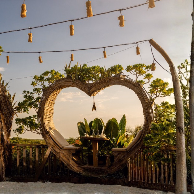 Two large heart-shaped wicker swings on the Karma Rooftop face lush greenery and the ocean, with string lights above and hanging decor in the center. The peaceful scene is framed by plants and bathed in soft sunset light.