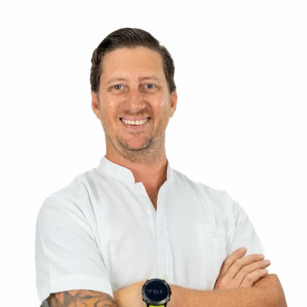 A smiling photographer with brown hair, in a white shirt and black watch, stands with arms crossed against a white background.