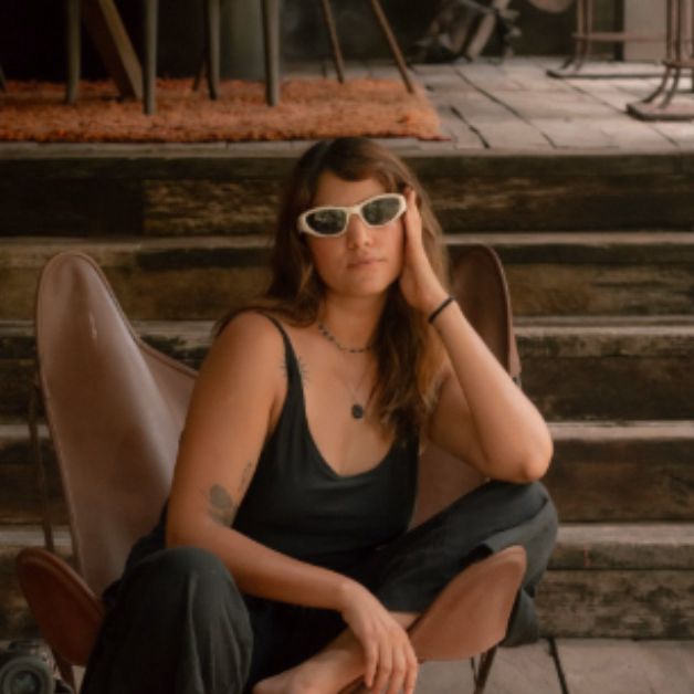 A woman in white sunglasses and a black jumpsuit sits casually in a chair, head resting on hand, stairs behind her.