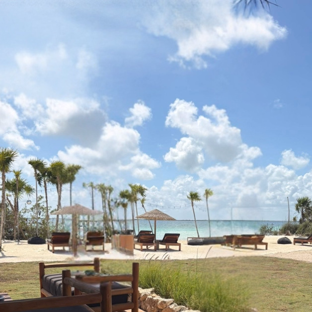 Wooden lounge chairs and umbrellas are arranged on the sandy beach at Sipalitos Lagoon Club, with palm trees and greenery nearby, overlooking a calm ocean under a partly cloudy blue sky.
