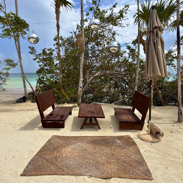 Wooden benches and a table on sand at Sipalitos Lagoon Club, trees and disco balls above, turquoise ocean in the background.
