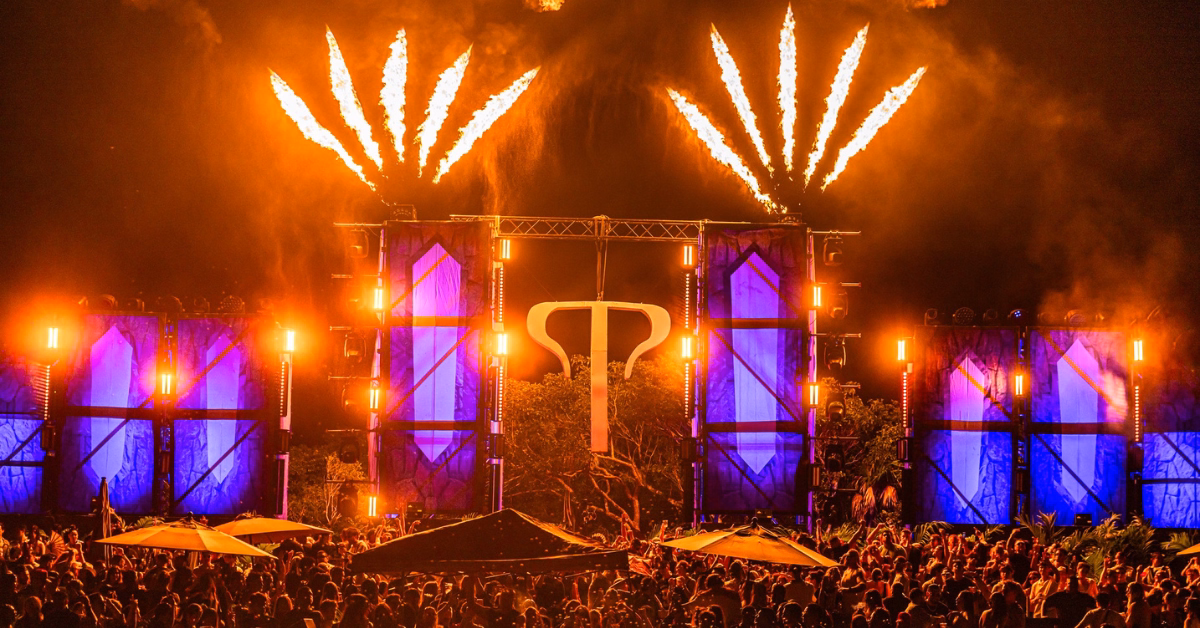A large crowd enjoys the nighttime tehmplo music festival, with a brightly lit stage featuring blue panels, orange lights, and tall bursts of fire shooting up above the stage.