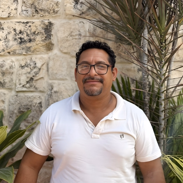 Smiling man with short curly hair, glasses, and a goatee stands by a stone wall and tropical plants in a white polo shirt.