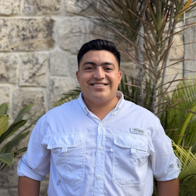 A man in a white button-up shirt stands smiling before a stone wall and green plants, looking professional and approachable.