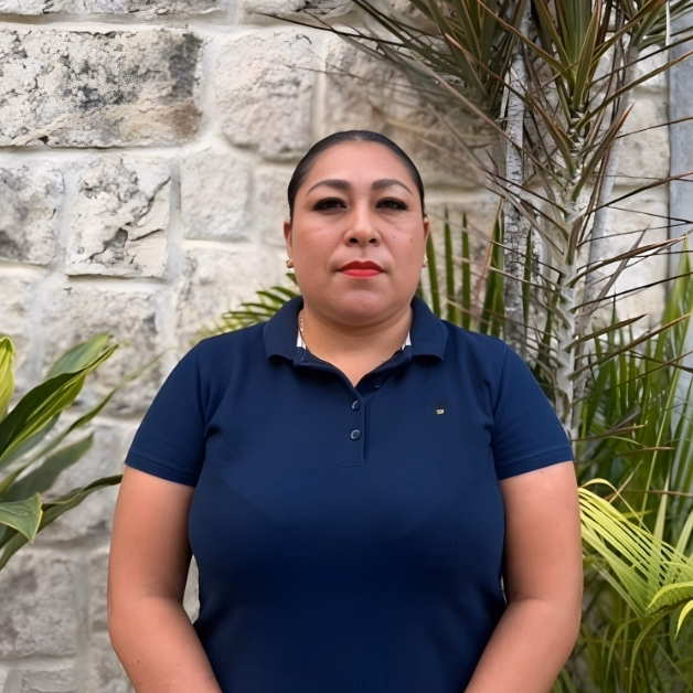 A woman with dark hair pulled back, in a navy polo shirt, stands before a stone wall and plants, facing the camera seriously.