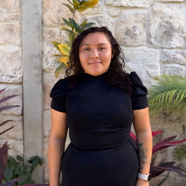 A woman with wavy dark hair in a black dress and white watch stands smiling by a stone wall with green and purple plants.