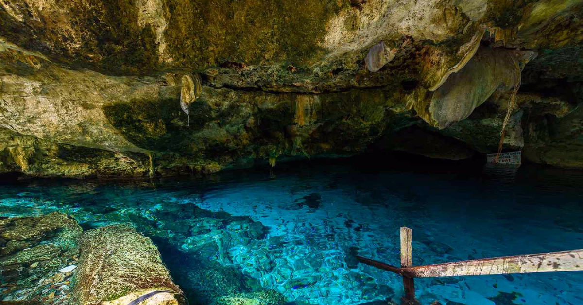 Turquoise water fills an underground cave with rocky, mossy ceilings and stalactites. Sunlight reveals rocks and a railing.