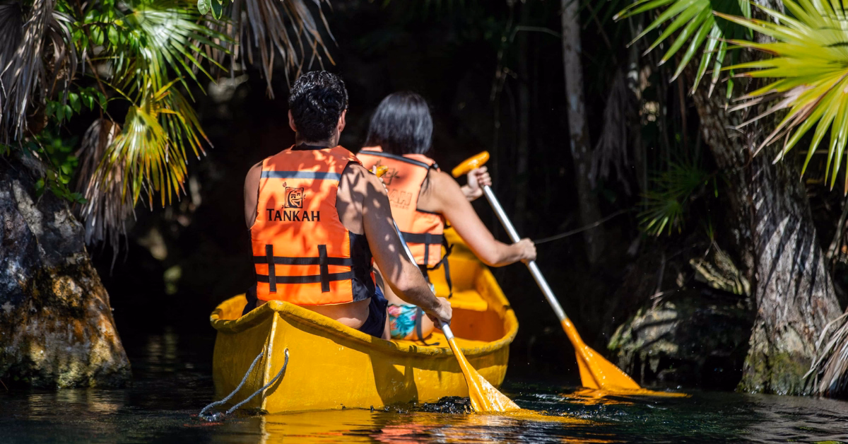 Two people in orange life vests paddle a yellow canoe through a narrow, lush tropical waterway surrounded by green plants.