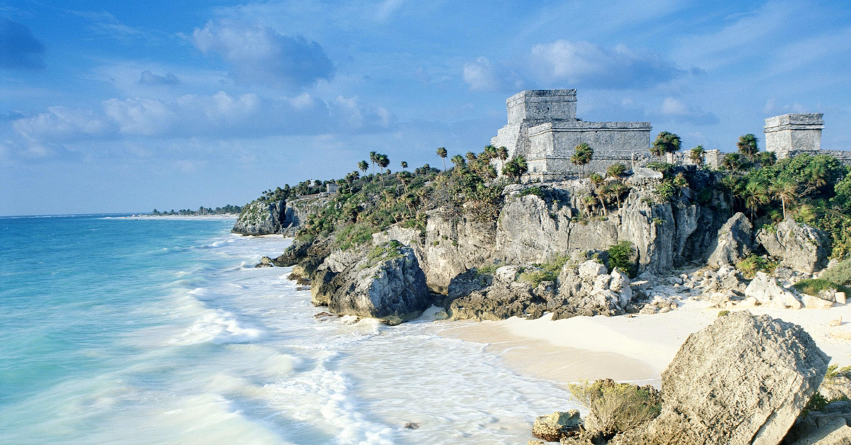 Mayan ruins sit on a rocky cliff above turquoise sea and white sand, palm trees dotted around under a partly cloudy sky.