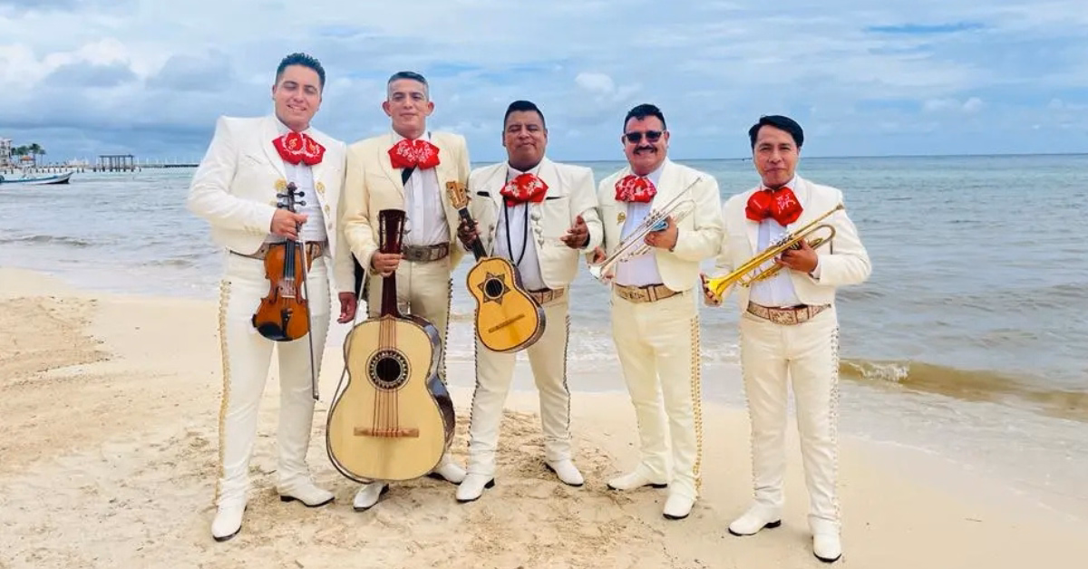 Five mariachi musicians in white suits with red bow ties pose with instruments on a sandy beach by the ocean under cloudy skies.