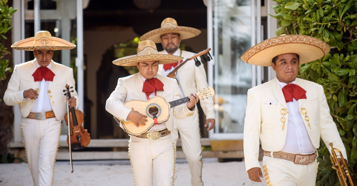 Four mariachi musicians in white suits and sombreros walk outside. One plays guitar, another has a violin. Plants and a doorway behind.