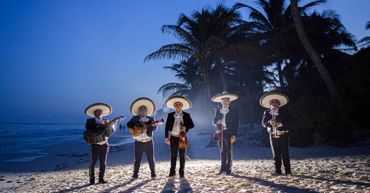 Five mariachi musicians in traditional outfits and sombreros play on a beach at dusk, with palm trees and the ocean behind them.