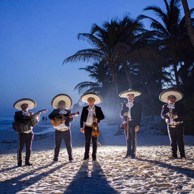 Five mariachi musicians in traditional outfits and sombreros stand on a sandy beach at dusk, ocean and palms behind them.
