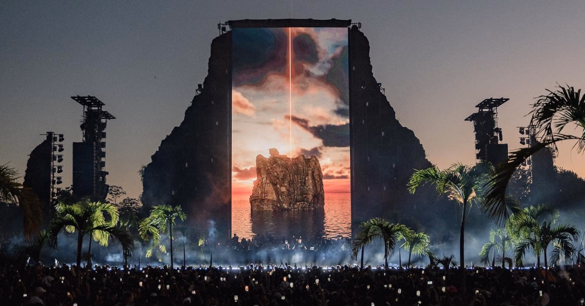 A large outdoor concert stage with a giant screen displaying a sunset over the ocean. Palm trees frame the stage at Zamna Music Festival, and a crowd is gathered, many holding up their phones as night falls.