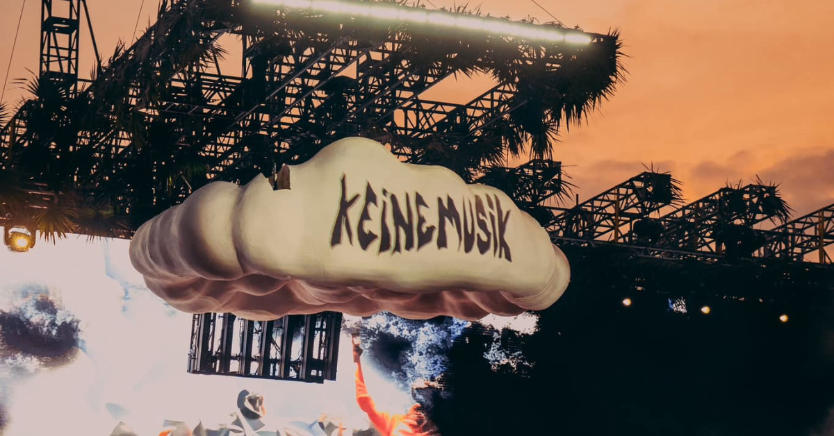 A large white cloud prop with KEINE MUSIK written on it hangs above a stage with lights and rigging, set against an orange sunset sky at the Zamna Music Festival.