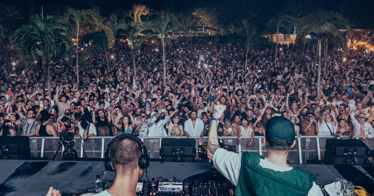 A large crowd enjoys the Zamna Music Festival at night, with two DJs performing on stage in front of palm trees. Many people in the audience have their hands raised and are taking photos.