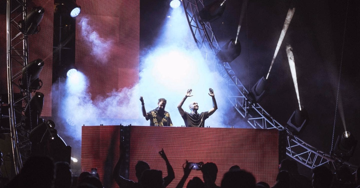 Two DJs perform onstage with their hands raised behind a large console at Zamna Music Festival, colorful lights and smoke effects surrounding them, while an energetic crowd dances and takes photos in the foreground.